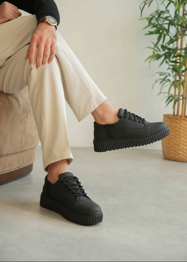Person wearing black chunky sneakers with beige pants on a light-colored floor.
